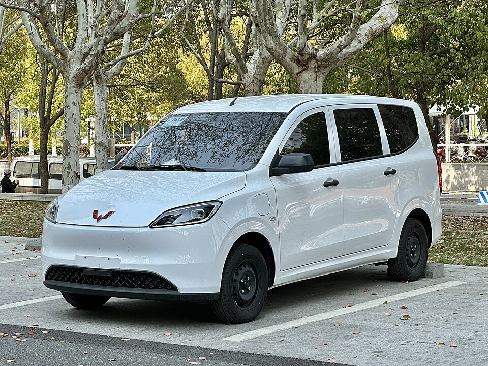Third generation Wuling Hongguang EV front quarter view in white paint. It is parked in a parking lot rear-in outdoors on a sunny evening with trees in the background.